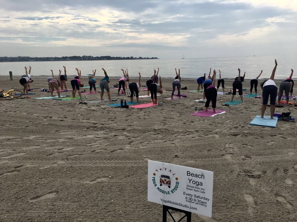 A Yoga Mobile Studio class on a beach, everyone reaching one arm overhead in a side stretch beside the water