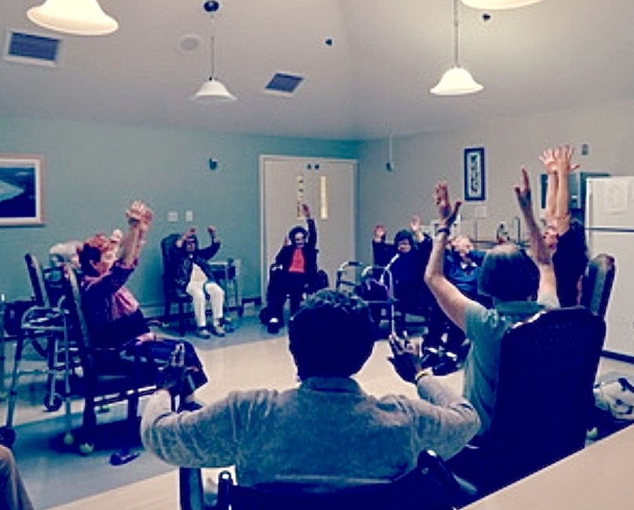 A seated group class in a bright common room, everyone reaching both arms overhead from their chairs, some with walkers beside them