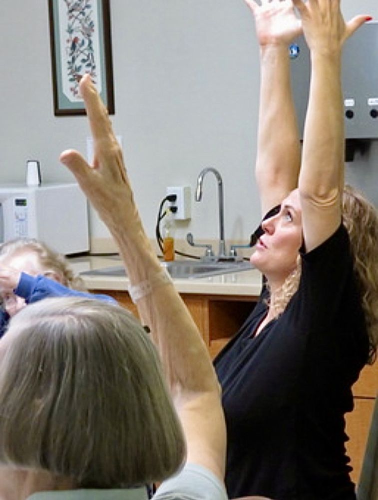 VictoriAnn guiding a seated class with both arms stretched high overhead, other participants' raised arms in the foreground