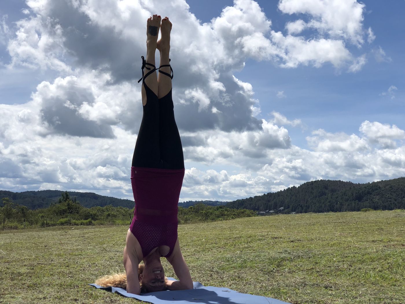 VictoriAnn in a headstand on a blue mat in a grassy field under big clouds, with green hills in the distance