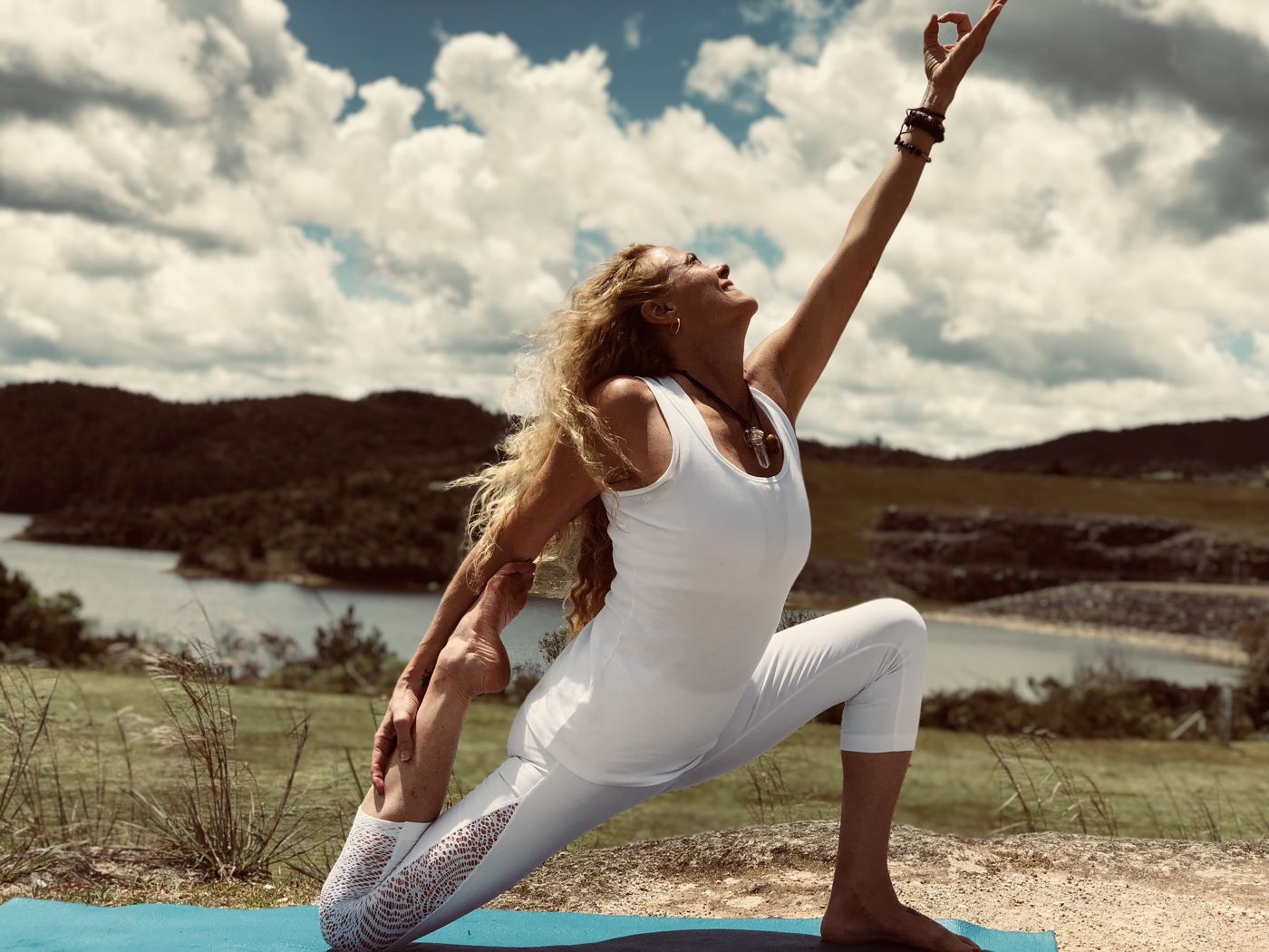 VictoriAnn in white in a low lunge on a blue mat, one arm reaching to the sky, a lake and hills behind