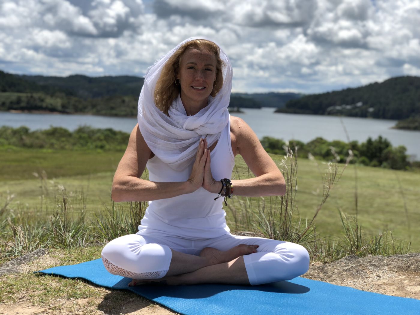 VictoriAnn seated outdoors in white with a light scarf over her hair, palms pressed together at her heart, a lake and hills behind her